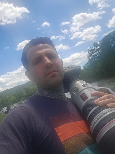 A selfie taken outdoors in Zimnitsa, Bulgaria, shows a man holding a professional camera with a large telephoto lens. The man has short dark hair and tattoos on his neck. He is wearing a t-shirt with orange, brown, and blue stripes. He is positioned with a view of green hills and trees in the background. The sky is visible, with scattered white clouds. The photo angle is tilted.