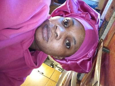 A close-up, vertically oriented photograph depicts an individual with dark skin, wearing a fuchsia-colored hooded sweatshirt and a matching fuchsia satin ruched head covering. A small metallic nose ring is visible on the subject's left nostril. The subject's eyes are open and directed towards the camera; the lips are slightly parted. The background, out of focus, indicates an indoor, domestic environment. Visible elements include a polished wooden surface with a lighter trim to the right of the subject and a light-colored floor, possibly tiled or hardwood, below. Other indistinct household items are vaguely discernible in the blurred background. The scene is located in Madinaty, Egypt.