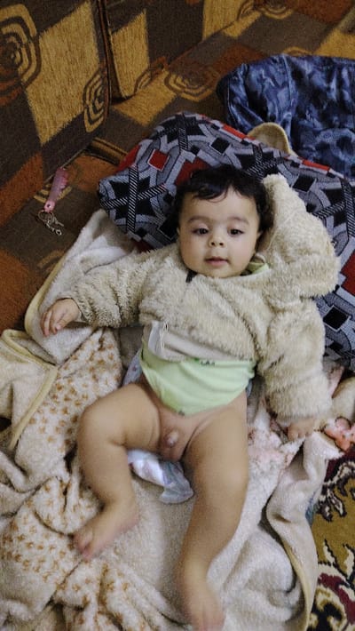 A baby lies on a patterned blanket, wearing a fuzzy jacket and a green undershirt over a diaper. The baby's legs are exposed. The background includes a patterned sofa and a decorative pillow. A pink comb-keychain rests near the baby's head. The lighting is bright and even, illuminating the scene.