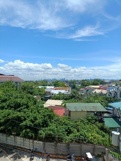 The image is a view from a rooftop overlooking a residential neighborhood in Parañaque, Philippines (latitude: 14.474, longitude: 121.008). The scene is dominated by lush green foliage, obscuring much of the cityscape in the distance. A concrete wall with a wire fence runs horizontally across the foreground.  A scattering of houses with various colored roofs are partially visible.  The sky is a clear, light blue with white clouds.  There are a few motorcycles parked on a paved area near the wall.  The distant cityscape is obscured by trees and clouds.  A large, white, industrial-looking structure is visible in the bottom right corner of the image.  The overall mood of the image is peaceful and serene.