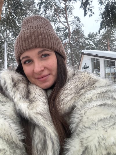 A young adult female is depicted in a head-and-shoulders portrait, positioned outdoors in a snowy environment. She is wearing a light brown ribbed knit beanie and a thick, multi-toned white and grey faux fur coat. Her long, dark brown hair is visible beneath her hat and draped over her shoulders. Small specks of snow are present on her hat, hair, and coat. She has fair skin, blue-green eyes, and a slight smile, looking directly at the viewer.

The background consists of snow-covered pine trees. To the subject's right, a white, modern-style building with large windows and a snow-laden roof is partially visible. A sign on the building displays the Cyrillic text "ТРАПЕЗНАЯ." This building is identified as the "Trapeznaya" cafe/dining hall located at the Ski Complex Kalinovo (Горнолыжный комплекс Калиново) in Kalinovo, Sverdlovsk Oblast, Russia. The scene conveys a cold, winter day with active snowfall.