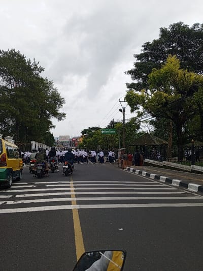 A street scene shows motorcycles and a yellow vehicle navigating a crosswalk. A large group of people, appearing to be students in uniforms, are crossing the street. Trees and a building with a gazebo are visible on the right side of the road. A street sign indicating "Istana Cipanas" is also present. The sky is overcast.