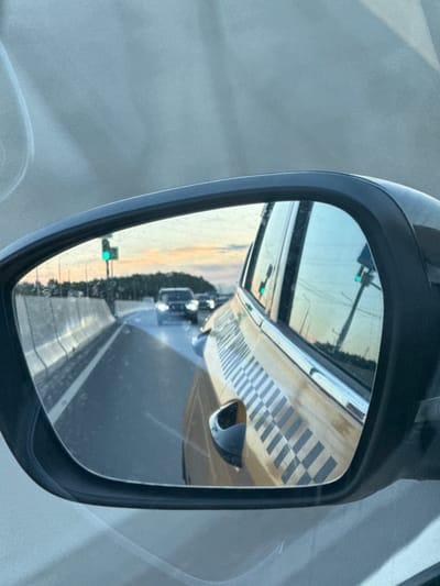 The image is a rear-view mirror reflection of a road scene in Borodino, Russia. The reflection shows a highway with a concrete barrier on the left and traffic in the distance, including cars and a traffic light displaying green. The sky is partly cloudy with hues of orange and yellow, suggesting it is either sunrise or sunset. The mirror itself is framed by a black car mirror housing, and part of a yellow car with a checkerboard pattern is visible in the foreground of the reflection. The overall environment appears to be urban or suburban.