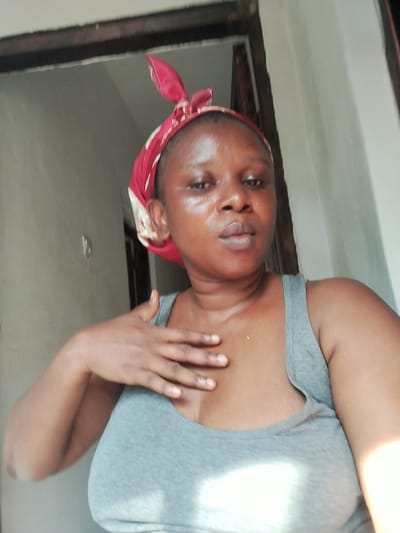 A dark-skinned individual, female, is depicted in a medium-shot selfie, gazing directly into the camera. She wears a grey tank top and a red and cream patterned head covering tied at the top of her head. Her left hand rests flat against her upper chest/sternum area. The individual's skin appears to have a sheen, particularly on the face. She is positioned indoors, with a light-colored wall visible behind her on the left and a dark wooden door frame or entrance on the right. The lighting suggests an indoor setting with ambient light possibly entering from the front or side. The specific location within the city of Abor, Nigeria, cannot be determined from the visual evidence present in the image.