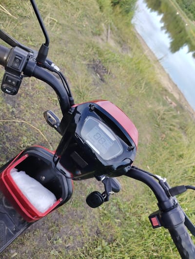 The image shows a close-up, eye-level view of the handlebars and dashboard of a red and black electric scooter. The scooter is parked outdoors on a grassy area next to a body of water.
**Subjects:** The primary subjects are the scooter's controls and dashboard. Visible are the handlebars, a rear-view mirror, a set of switches on the left handlebar including what appears to be a headlight control and turn signals, and a horn button. The dashboard displays a digital screen showing speed and battery level, with text "Dreams of Science" and "Km/h" visible. A key is inserted into an ignition on the right side of the dashboard. A red storage compartment is open, revealing white padding inside.
**Scene/Setting:** The setting is outdoors, with a focus on the scooter's front. The background shows a grassy field, a dirt path, and a body of water reflecting trees and the sky. The overall impression is one of a natural, possibly rural environment.
**Activity/Event:** No active event is taking place; the scooter appears to be parked.
**Notable Details:** The time of day is not clearly indicated, but the lighting suggests it could be daytime. The weather appears to be overcast or cloudy, judging by the reflection in the water. The location context is provided as Заямнаўскі сельскі Савет, Belarus, which suggests a rural area in Belarus. There are no people visible in the image.
**Visible Text:**
* Dreams of Science
* Km/h