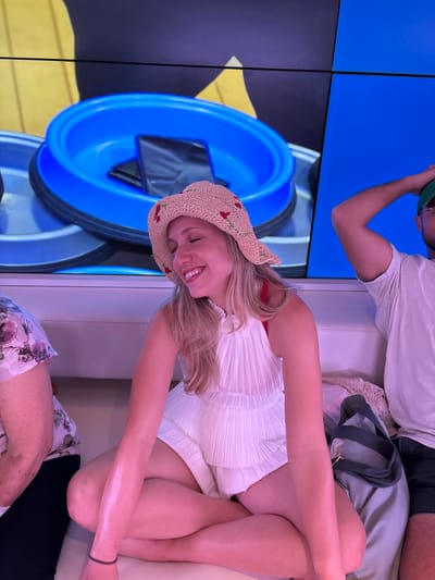 A young woman wearing a white dress and a straw hat adorned with red cherries sits cross-legged on a white couch. She smiles with her eyes closed. Behind her is a large screen displaying a blue dog bowl with a black phone inside. To her left sits a person in a floral top, and to her right sits a man with a green hat. A gray bag rests between the woman and the man.