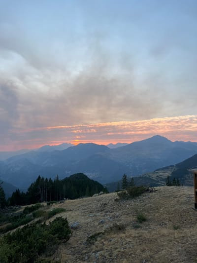This is a photograph of a mountain landscape at dusk. In the foreground, dry, golden grass covers a sloping hill dotted with scattered rocks and low-lying green shrubs. A line of dark green coniferous trees stands on a lower ridge to the left. In the middle ground, layers of progressively lighter blue mountains recede into the distance. The setting sun, a bright orange disc, is visible just above the horizon, casting a warm glow on the clouds. The sky transitions from a pale blue at the top to hues of pink and orange near the horizon, with scattered grey clouds. The scene appears to be in a rural, mountainous area, likely in the vicinity of San Juan de Plan/San Chuan de Plan, Spain, given the distinctive mountain ranges and vegetation typical of the Pyrenees. There are no people or animals visible in the image.