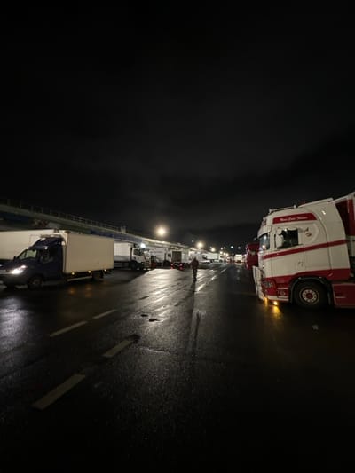 A nighttime scene depicts a long line of parked semi-trucks stretching into the distance on a wet, paved surface. Streetlights cast a hazy glow, reflecting off the slick ground. Most of the vehicles are white, with one prominent red and white truck parked near the top of the frame. A single figure appears to be walking alongside the trucks. The overall atmosphere is dark and somewhat desolate.