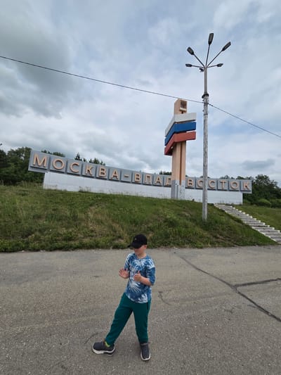 A young boy wearing a tie-dye shirt, green pants, a black baseball cap, and sneakers stands on an asphalt road. He is looking to his left with his hands raised as if clapping or gesturing. In the background, a large sign with Cyrillic lettering that reads "МОСКВА-ВЛАДИВОСТОК" is situated on a grassy hill. The sign is accented with red, white, and blue stripes. A tall lamp post with multiple lights stands next to the sign. The sky is overcast with gray clouds. Stairs are visible on the right side of the hill. The location is Nikolaevka, Russia, a town known for being the starting point of the Trans-Siberian Highway.