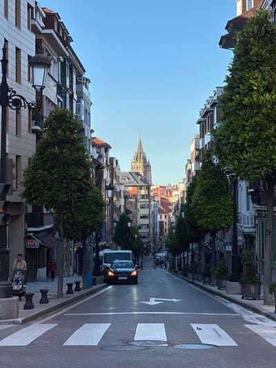 The image captures a street scene in Oviedo, Spain, likely on Calle Uría, a prominent avenue known for its elegant architecture and lively atmosphere. The perspective is from a pedestrian crossing, looking down a slight incline towards a distant cathedral spire, a recognizable landmark of the city.

On either side of the street, imposing buildings with classic European architecture rise. Their facades are a mix of light-colored stone or stucco and darker brick, punctuated by numerous windows and balconies. Several manicured trees, with their dense green foliage, are strategically placed along the sidewalks, adding a touch of nature to the urban landscape. Intricate street lamps adorn the pavements, hinting at the city's historical character.

A black Mercedes sedan is the most prominent vehicle on the road, its headlights illuminated. Other vehicles are visible further down the street, suggesting moderate traffic. Pedestrians are present but not in large numbers. On the left side of the frame, a woman is seen pushing a stroller, while another individual walks further down the sidewalk. The street itself is paved with asphalt and marked with white lines for traffic lanes and a prominent pedestrian crossing.

The sky above is a clear, pale blue, indicating fair weather and daylight, likely late afternoon or early morning given the lighting. The overall impression is one of a well-maintained and picturesque European city street.