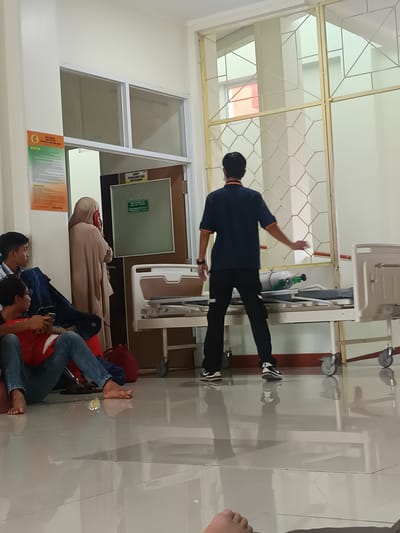 The image depicts an indoor scene, possibly a hospital waiting area in Karangtengah, Indonesia. In the foreground, three young men are seated on the floor, with one of them leaning against a red bag. They are casually dressed in t-shirts and jeans. To the left, a woman in a beige hijab is standing and appears to be talking on her phone. She is near a doorway labeled "UNIT PERAWATAN INTENSIF," indicating an Intensive Care Unit. In the middle ground, a person in a dark blue uniform is standing next to a hospital bed that is partially obscured by a metal grate. An oxygen tank is visible on the bed. The floor is tiled and reflective, showing the reflection of the people and objects in the room. The overall lighting suggests an indoor, possibly fluorescent, illumination.