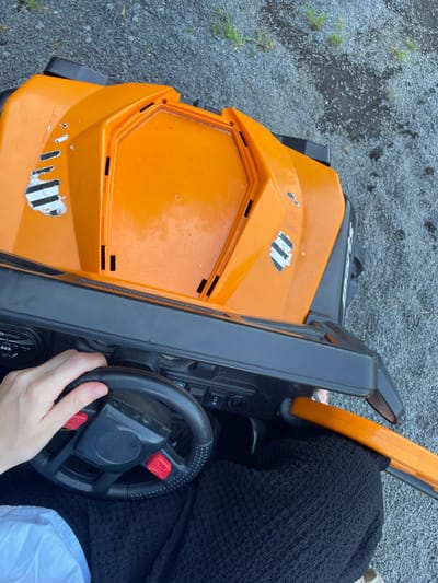 An overhead, close-up shot captures a person's hand gripping the steering wheel of an orange toy vehicle. The vehicle is positioned on a gravel surface, likely a driveway or outdoor play area. The toy car's body is predominantly orange with black accents and features some wear and tear, including chipped paint and what appear to be remnants of racing stripes. A black dashboard with various buttons and a small screen is visible beneath the steering wheel. The person's hand is casually resting on the black steering wheel, with fingers slightly curled around it. Their attire consists of black textured fabric visible around the vehicle's interior, and a portion of a white garment is also visible. The background consists of small, gray gravel and sparse green vegetation. The location is identified as Beltershain, Germany.