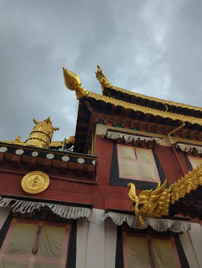 This image shows the exterior of a building, likely a temple, with ornate gold decorations. The building has red walls and a dark roofline adorned with intricate gold trim and dragon-like sculptures. A large, golden finial, shaped like a lotus bud, stands prominently against a cloudy grey sky. Below the roofline, several windows are visible, covered with light-colored fabric. A circular golden emblem featuring a seated figure is mounted on the red wall. This building is located in the city of Shangri-La, China, a region known for its Tibetan Buddhist monasteries.