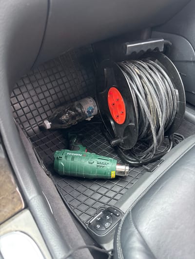 The image shows the interior of a vehicle, likely the footwell of a passenger seat. A black rubber floor mat covers the floor. On the mat, there is a large black cord reel with a red section containing three electrical outlets. A green heat gun, labeled "PARKSIDE," is positioned in front of the cord reel. To the left of the cord reel and heat gun is a clear plastic bottle, possibly containing liquid, with a white cap. The interior of the car appears to be dark-colored, with leather seats visible. The background suggests the car is stationary. There are no people present in the image. The scene is forensic in nature, providing a detailed snapshot of objects within the vehicle. The location provided, Yoakim Gruevo, Bulgaria, is noted as the context for this scene.