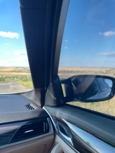 The image is a first-person perspective from inside a car, looking out the passenger side window. The view is of a rural landscape under a bright blue sky with scattered white clouds. In the foreground, the car's interior is visible, including a portion of the dashboard with an air vent and a glossy trim piece with a rainbow-like iridescence. The side mirror is also in view, reflecting the sky and the car's interior. The exterior scene shows a winding road leading through a grassy, somewhat arid landscape with scattered trees and buildings in the distance. The location is identified as being in Malynivka, Ukraine.
