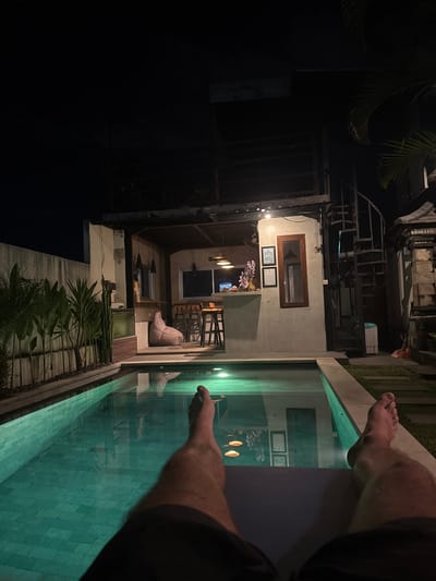 The image depicts an outdoor area at night in Munggu, Indonesia. A pool is visible in the foreground, with the feet of a person dangling over the edge. The pool's water appears to be illuminated, and the pool itself is lined with tiles. Behind the pool is a building or structure featuring a window and open-air dining or lounge area with a beanbag chair and bar stools. Above the building appears to be a skeletal metal structure. The left side of the image is dominated by darkness, possibly indicating a dark structure or dense foliage. Plant life is visible near the base of the building.