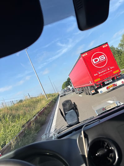 This image captures a view from inside a vehicle, looking through the windshield. The scene depicts a highway with moderate traffic under a clear blue sky with sparse clouds. A large red semi-trailer truck with "DS TRANSPORT" and "DE SMEDT & ZN." written on its side dominates the right side of the frame. A white van is visible in the distance to the left, and other vehicles are present in the traffic flow. The roadside on the left features a grassy embankment with a metal guardrail. A tall lamppost stands next to the highway. In the foreground, parts of the vehicle's interior are visible, including a dashboard with a device mounted on it and a cup holder. The overall impression is a snapshot of a typical highway scene in daylight. The provided context indicates the location is the city of Vilvoorde, Belgium.