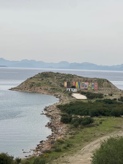 The image shows an aerial, sideways view of a promontory extending into the sea, likely taken in Datça, Turkey. The landscape is dominated by the shoreline, with the clear water stretching into the distance where hazy mountains fade into the sky. The promontory itself is covered in sparse vegetation, dirt, and rocks. At the end of the promontory, closest to the viewer, is a series of eight brightly painted panels displaying various symbols. Behind the panels are a few small white buildings. A dirt path runs along the top of the promontory on the right side of the image, bordering the vegetation. The sky is overcast, contributing to the muted color palette.