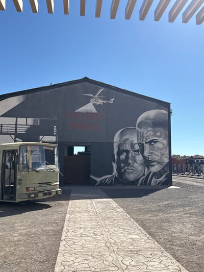 The image displays an outdoor scene at a film studio, likely Atlas Film Studios or CLA Studios, located in Ouarzazate, Morocco, under a clear blue sky and bright sunlight. The central feature is a dark grey, single-story building with a prominent mural covering its facade. The mural depicts a light-colored helicopter projecting a searchlight beam, the red text "PRISON BREAK," and two stylized monochrome portraits of men with shaved heads; one figure, positioned left, appears to be smoking, while the other, on the right, has a stern expression.
To the left of the building, an olive-green, older model bus is parked, facing forward. It appears to be a prop, with its front bumper, grille, and headlights visible, and possible missing side elements. A light-colored, paved pathway with a repeating geometric pattern leads directly from the foreground towards a dark, rectangular opening in the building, functioning as an entrance. The surrounding ground is composed of dark, loose gravel or dirt. Above, a series of parallel, light-colored horizontal beams partially frame the top of the image. Distant red and dark structures are faintly visible on the far right. No individuals are present in the immediate scene.