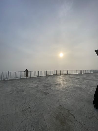 A lone person stands at a white railing overlooking a foggy expanse of water, likely Lake Baikal, near the city of Listvyanka, Russia. The sun is a diffused bright orb in the hazy sky. The foreground is a grey concrete patio with a rough, textured surface, appearing wet in places, reflecting the low light. The railing runs horizontally across the midground, separating the patio from the water. The figure, seen from behind, is wearing light-colored shorts and a light-colored shirt. Their posture suggests contemplation or observation of the water and the fog. To the far right, a dark, angular object, possibly part of a structure, juts into the frame from the edge of the image. The overall atmosphere is serene and slightly melancholic due to the fog and muted light.