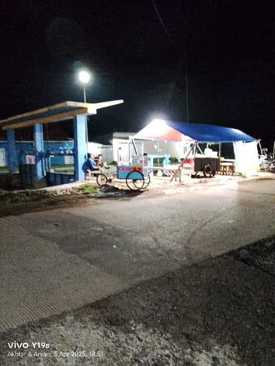 The image shows a street in Kecamatan Kroya, Indonesia, at night, illuminated by streetlights. There is a small, brightly lit food stall with a blue tent, sitting on the right side of the street. A person is standing in front of the stall. Another person is sitting on a bench, positioned under a blue canopy on the left side of the road. In the background, there are some buildings and trees. The ground is paved with asphalt and is partially covered with grass. The scene appears to be a street in an urban or suburban area of Kecamatan Kroya, Indonesia. The location's latitude is -6.422 and longitude is 107.996.