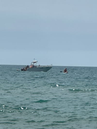 The image depicts a body of water, likely the sea, under a pale blue sky. In the middle ground, a white motorboat with a canopy is visible, carrying several people. To the right of the boat, a person is sitting on a paddleboard, holding a paddle. The water is choppy with small waves, and there are glints of sunlight on the surface. The overall scene suggests a recreational activity taking place on the water. Given the context of Vityazevo, Russia, this scene is likely at the Black Sea coast.