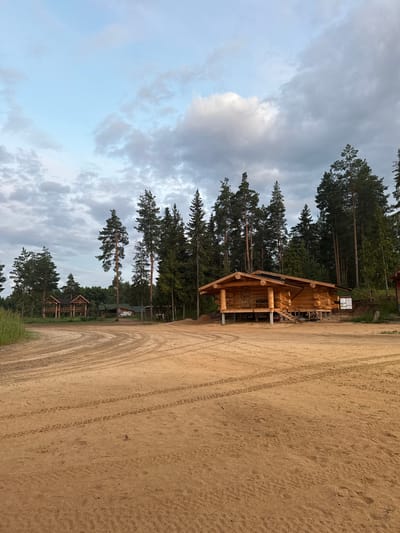 The image depicts an outdoor scene in Сосновка, Russia, likely during the daytime with overcast skies. The foreground is dominated by a sandy, unpaved path with tire tracks clearly visible. To the left of the path, a dense forest of tall pine trees stretches into the distance. Among the trees, several wooden structures are visible.

The most prominent structure is a partially constructed wooden cabin, made of logs, situated on stilts. Its roof is partly visible, and construction materials and scaffolding suggest ongoing work. Further back, nestled within the trees, are other smaller wooden buildings that appear to be cabins or outbuildings, some with visible roofs and windows.

The overall impression is of a rural or semi-rural setting, possibly a recreational area or a developing campsite. The weather appears cloudy, creating a diffused light. The scene is quiet and still, with no people or animals immediately visible, emphasizing the natural environment and the man-made structures within it. A small sign with text is partially visible near the main cabin but is not legible.