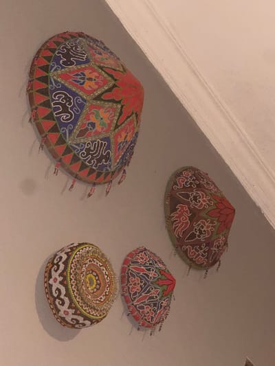 The image shows four decorative, conical hats hanging on a light-colored wall. The hats are adorned with intricate beadwork in various patterns and colors, including red, blue, green, black, and yellow. One hat in the upper left is primarily red, blue, and black with floral and abstract designs. To its right and slightly lower is a hat with a brown base and red and white patterns. In the lower left, there is a round, flattened hat with a circular, multi-colored design. Adjacent to it and slightly to the right is another conical hat with red, blue, and green patterns. The hats appear to be of traditional design, possibly from Southeast Asia. The environment is indoors, with the wall showing a slight texture and a subtle shadow cast by the hats. The lighting suggests an indoor setting, potentially a home or a cultural space. The image does not provide any information about the city of Abiansemal, Indonesia, or any specific context related to its location.