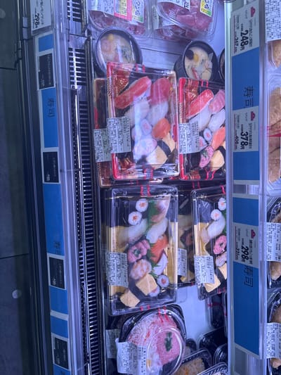 The image shows a refrigerated display case in a grocery store. The case contains various sushi offerings, including a variety of nigiri (hand-pressed sushi rice topped with seafood and vegetables) and maki (sushi rolls). The sushi is presented in clear plastic containers with white lids. The containers are arranged in two rows with the sushi in the front row and other food items such as meat, soup, and a bowl of rice with toppings in the back row. Each container of sushi has a price tag attached that shows the price in Japanese Yen. The price tags also contain Japanese characters indicating the type of sushi. The bottom row of the display case has a blue sign that reads "ari" which means "available" in Japanese. The display case is located in a grocery store in Minato City, Japan, at latitude 35.67 and longitude 139.716. The image was taken from the front of the display case, looking at the sushi at eye level.