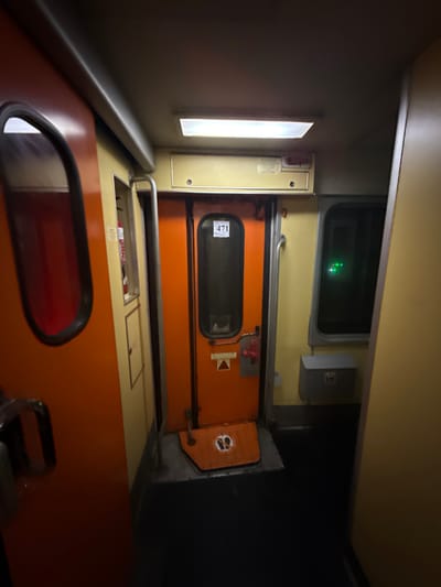 Inside a train car, an orange door with a window labeled "471" is visible. Overhead, a rectangular window shows green lights. Next to the door, an emergency exit handle is visible. Below, an orange seat with a red window and metal armrest is present. The train car's wall is a pale yellow.
