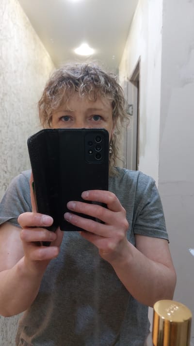 A woman with blonde, curly hair is taking a mirror selfie. She is wearing a grey t-shirt and holding a black smartphone in front of her face, obscuring her mouth and nose. Her eyes are visible, and they are blue. She is wearing dangling earrings. The background appears to be a bathroom or hallway with textured walls and a door. A gold-capped bottle is partially visible in the bottom right corner of the image. The lighting suggests an indoor environment. The location of Зелёный, Russia, is not discernible from the image.