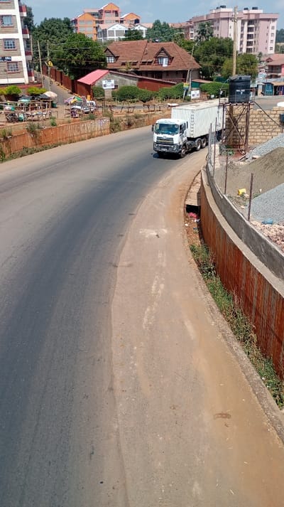A wide-angle, elevated daylight image captures a section of a road and surrounding urban landscape in Kinoo ward, Kenya.
**Road and Immediate Surroundings:** The primary feature is a two-lane asphalt road, partially shaded in the foreground, curving towards the left as it recedes into the background. A lighter-colored dirt/gravel shoulder parallels the road on the right. Adjacent to this shoulder, a retaining wall composed of rusted corrugated metal sheets topped with concrete runs along the right side of the frame. Beyond this wall, construction is evident, with a pile of grey aggregate (gravel) and a partially constructed wall of grey cinder blocks. A black plastic "VELTUS 1000L" water tank is mounted on a metal scaffold structure directly above the aggregate pile. A narrow dirt path and drainage ditch, containing some refuse, separate the road from the retaining wall.
**Left Side of the Road:** Elevated above the main road on the left, an unpaved area features several informal market stalls with wooden frames and coverings such as blue tarps and a yellow umbrella. At least two individuals are discernible near these stalls, one beneath the blue tarp and another nearby. Behind these stalls, a two-story residential building with "WEST HILL" visible on its facade stands. Further back, a distinct building with a red tiled roof and multiple dormer windows is visible, along with a smaller adjacent structure with a red corrugated roof. Utility poles and power lines are present.
**Background:** The background reveals a panorama of multi-story buildings, characteristic of urban development. These structures display varied architectural styles and colors, with some featuring reddish-orange roofs and others having lighter facades. Green foliage and trees are interspersed among the buildings, indicating a mix of natural and built environments under a clear blue sky.
**Vehicles and People:** A white UD Nissan tractor-trailer unit, labeled "KAVANSOWA," is captured in motion on the road, traveling away from the viewer. It is pulling a white, ribbed shipping container. The two individuals near the market stalls are the only discernible people.
**Overall Environment:** The scene depicts an active urban area undergoing development, characterized by a mix of established buildings, informal commerce, and ongoing construction.
