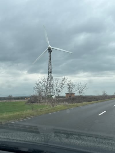 The image displays an overcast, rural scene near Gorno Aleksandrovo, Bulgaria. A prominent wind turbine, featuring three white, stationary blades atop a metallic lattice tower, occupies the left-center of the frame. To the left of the turbine, a vibrant green field extends, bordered in the distance by a long, dark horizontal structure, possibly a barrier or solar array, with a yellow object visible nearby. A cluster of bare, deciduous trees and shrubs is situated between the field and a wet asphalt road. Behind these trees, to the right of the turbine, stands a small, square, single-story building constructed of reddish-brown brick with a dark roof. The asphalt road, running from the lower right toward the upper left, is dark and reflective, suggesting wet conditions. It features a solid white line on its right edge and a broken white line partially visible as a center marker. Distant, hazy mountains are visible on the horizon beneath the grey, cloudy sky. The photograph was captured from within a vehicle, with the dashboard or lower windshield visible in the foreground. No individuals are present in the scene.