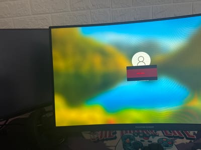 The image shows a computer setup. There are two monitors, one to the left which is mostly out of view, and a curved monitor in the center displaying the Windows login screen. A red error message overlay says "DisplayPort." The monitor's bottom bezel reads "PHANTOM GAMING." Below the monitor is a patterned desk mat featuring what appears to be a gaming controller design. The background includes a white brick wall. There is no information related to Saedinenie, Bulgaria.