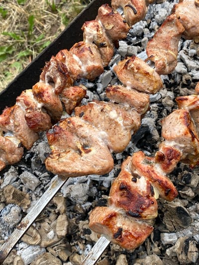 This media file shows skewers of meat cooking over hot coals. The meat appears to be pork, cut into bite-sized pieces and threaded onto metal skewers. The skewers are arranged over a bed of glowing charcoal, with some pieces of meat showing browning and charring from the heat. The background consists of the charcoal grill and some dry grass and green foliage, suggesting an outdoor setting. The lighting and the appearance of the coals indicate that it is likely daytime, possibly late afternoon or early evening, as the coals are well-lit. There are no people visible in the image. The scene depicts the process of grilling, likely a barbecue or a preparation of shashlik, a popular dish in Eastern Europe, which aligns with the provided location context of Velykyi Bychkiv, Ukraine. No text is visible in the image.