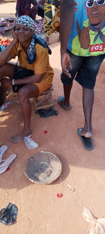 This media file depicts an outdoor market scene in Ketu Adieowe, Nigeria, characterized by its dusty, reddish-brown ground and bright natural light, suggesting daytime.

In the foreground, a woman is seated on a small wooden stool on the left. She wears a mustard yellow short-sleeved top and dark shorts. Her head is covered with a white headscarf featuring a dark pattern, and a black or dark blue scarf is tied over it. She has a subtle smile on her face and rests her chin on her left hand, appearing relaxed. A small nose piercing is visible. A black shoulder bag is slung across her body. Her feet are bare, and a pair of white flip-flops are visible on the ground near her.

To her right, partially in the frame, stands a man. He wears a light blue t-shirt adorned with a vibrant graphic of a person with pink hair and white sunglasses, making a hand gesture. The text "FOS" is visible on the t-shirt. He is also wearing dark shorts. His right foot is clad in a dark slipper, while his left leg and foot are bare.

In the background, out of focus, are various market items, including baskets piled high with red onions and tomatoes. Other blurred figures of people are also present, one dressed in a dark purple or maroon outfit sitting further back.

The ground around the subjects is dusty and sparsely littered with items such as a round, woven straw mat or basket, a white plastic bag, a black plastic bag, and a red bottle cap. The overall scene suggests a casual, everyday market environment.