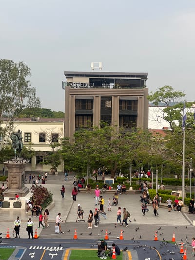 The image shows Plaza Gerardo Barrios in San Salvador, El Salvador. A crowd of people are gathered. The CAFÉ DE DON PEDRO building is visible. Also visible is the statue of Gerardo Barrios. There are orange cones along the street, and pigeons scattered on the ground.