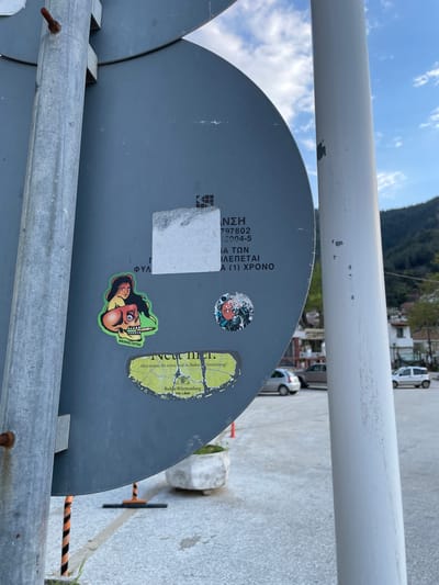 The image shows a close-up view of a gray information sign in the city of Skala Potamias, Greece. The sign has some Greek text that reads “ΝΣΗ 797802 16904-5 ΙΑ ΤΩΝ ΒΛΕΠΕΤΑΙ Α (1) ΧΡΟΝΟ," and is attached to a metal structure. Various stickers adorn the sign, including one of a stylized skull with the Instagram handle "@IRONSKI.TATTOO" at the bottom, one of a map that includes the text "Aber waren Sie schon mal in Baden-Württemberg? Baden-Württemberg THE LAND”, and another small, round sticker. The background shows a blurred street scene with parked cars and a glimpse of a mountain.