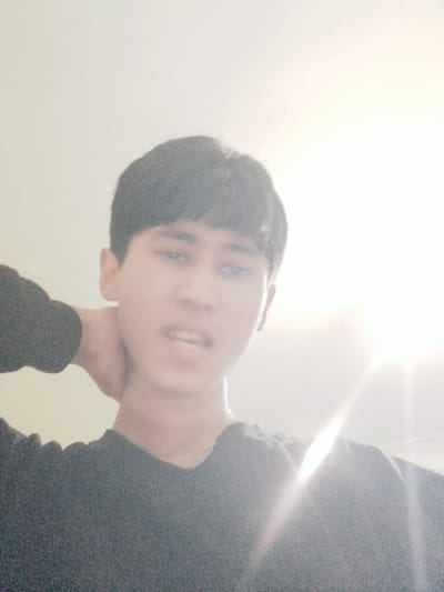 A young person is depicted in a close-up, highly overexposed photograph. The image is oriented sideways, with the person's head appearing on the left side of the frame and their body extending to the right. The person has short, dark hair, styled to cover their forehead. Their face is largely in shadow or underexposed compared to the extremely bright background, but their eyes, which appear dark brown or black, are visible and looking towards the viewer. Their mouth is slightly open, revealing teeth.
The person is wearing a dark-colored, possibly black, long-sleeved top or sweater with visible texture on the fabric. One hand is placed behind their head or neck, giving the impression of reclining or resting.
The background is almost entirely blown out by a very strong, bright light source, which appears as a large, intense white glow in the upper right quadrant of the image. This light source creates a lens flare effect and makes most of the background indistinguishable, appearing as a plain, bright white expanse. The overall lighting is very harsh, causing significant overexposure and a loss of detail in the brighter areas. No text is visible in the image.