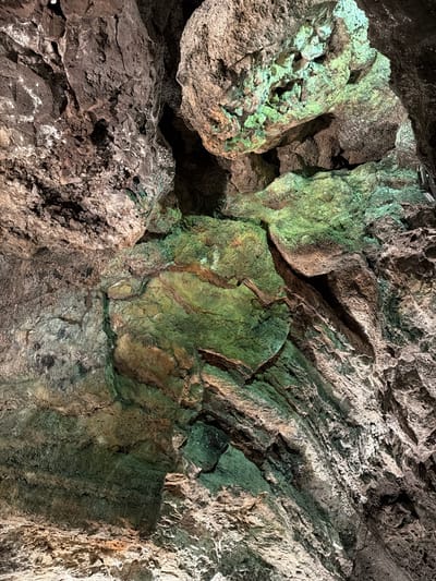 The image shows a close-up of rock formations inside Cueva de los Verdes, a lava tube located in Haría, Lanzarote, Spain. The rocks are various shades of grey, brown, and green, with rough, uneven surfaces. Some areas are lit with artificial green light, highlighting textures and irregularities. The composition fills the frame, giving a sense of depth and complexity to the geological features.