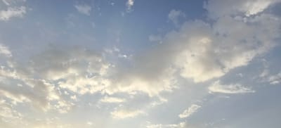 The image depicts a sky filled with scattered clouds of varying densities against a pale blue backdrop. The sunlight appears to be diffused, creating soft highlights on some of the cloud formations. The overall impression is that of a calm, perhaps early morning or late afternoon sky. There are no people, animals, or man-made objects visible in the frame. The scene is a natural environment, specifically the sky above Margilan City, Uzbekistan, as indicated by the provided context.