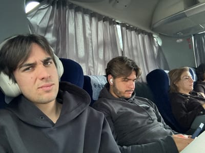 A photo shows three people and part of a fourth, seated on what appears to be an airplane. The individual nearest the camera is a young man wearing large headphones and a dark grey zip-up jacket. Next to him is another man in a grey hoodie and what looks like an earbud in his ear. Seated further back is an older woman in a dark jacket, and a portion of another person’s curly hair. The background consists of window curtains. The photograph seems to be rotated counter-clockwise.