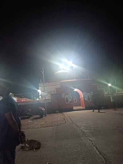 A nighttime outdoor scene depicts a red building with a domed roof, possibly a temple or mosque, illuminated by bright lights. The building features an arched entrance and is adorned with framed images on its facade. A person stands to the left, partially obscuring the view of the building's interior. To the right of the building, another person walks by. In the foreground, a dog is lying on the ground. Two motorcycles are parked on the paved area in front of the building. The lighting creates strong glare and lens flare in the upper portion of the image. The scene appears to be located in Bagerhat, Bangladesh.