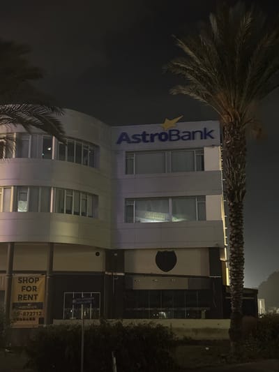 The image, captured at night, presents a multi-story commercial building featuring a modern design with a curved facade. The setting is an urban environment, specifically in Egkomi Lefkosias, Cyprus. Several tall palm trees frame the building, their fronds visible against the dark night sky.

The primary subject is the building itself, prominently featuring a large, illuminated sign on its exterior that reads "AstroBank" in blue capital letters, accompanied by a yellow star logo. The building has numerous rectangular windows across its floors, some of which show internal lighting and reflections. In one upper-story window, a reversed reflection of text, possibly "light2M," is discernible.

Adjacent to the AstroBank building, to its right, there is a darker, less illuminated structure. This structure displays a large, dark circular emblem or logo on its facade. Below this, a yellow banner is conspicuously hung, advertising a commercial space. The text on this banner clearly states "SHOP FOR RENT," followed by "Astrobank" and a contact phone number, "22-872712." Another smaller sign, partially obscured, is also visible near the banner.

The scene conveys a quiet, late-night atmosphere; no individuals or active events are present. The clear dark sky and illuminated signs confirm the time of day.