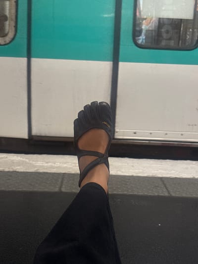 A person is standing on a metro platform, wearing black pants and a black Vibram FiveFingers shoe. The platform is tiled in gray. Behind the person, there is a train with a green and white exterior. The train is likely part of the Paris Metro system. The location is likely near a station in Clichy, France based on the visible exterior color scheme of the train that is common for the Parisian metro lines.