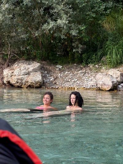 The image shows two young women swimming in a clear, shallow body of water. The woman on the left has long, dark hair and is looking towards the camera. The woman on the right has shorter, curly hair and is smiling. Both are wearing swimsuits. The water is surrounded by lush green vegetation on the left and a rocky shoreline. The water is clear and reflects the sunlight, creating a shimmering effect. The scene appears to be a natural swimming spot, possibly a river or a lake. The overall mood is relaxed and joyful, suggesting a warm, sunny day. The location is Spiljani, Bosnia and Herzegovina. There is no visible text in the image.