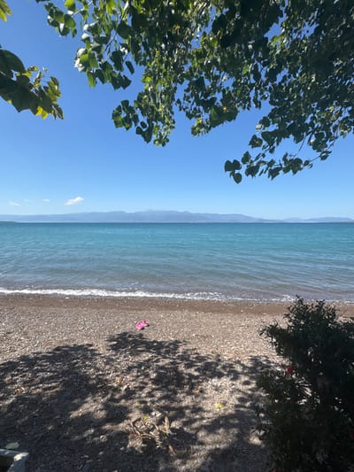 A serene view of a pebbled beach facing a calm, turquoise sea under a clear blue sky. In the foreground, sunlight filters through the leaves of a tree, casting dappled shadows on the beach. A pair of pink flip-flops rests on the pebbles near the waterline. In the distance, a mountain range forms a hazy horizon line. The scene evokes a peaceful summer day in Greece.