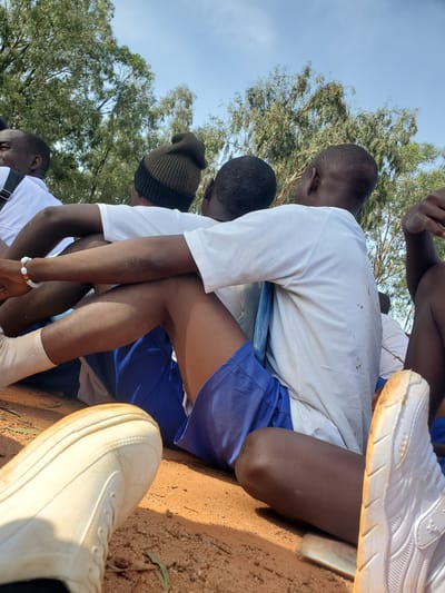 This media file depicts a group of young men, likely students, sitting outdoors on a sunny day. The scene is set in a rural or semi-rural location in Barakin Rafin Gora, Nigeria, with a backdrop of trees and a clear blue sky. The individuals are wearing casual attire, including white t-shirts and blue shorts, with one person wearing a dark beanie.

The subjects appear to be engaged in a moment of rest or anticipation, possibly before or after an activity. Their postures suggest a relaxed atmosphere. Notable details include the worn white sneakers visible in the foreground, indicating the practicality of their footwear. The ground appears to be dirt, further suggesting an outdoor, possibly recreational or educational setting. The lighting suggests it is daytime, likely mid-morning or afternoon. The overall mood appears casual and communal. There is no visible text in the image.