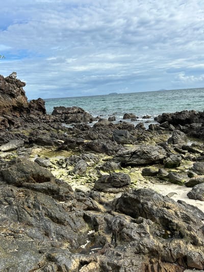 This is a photograph of a rocky coastline with the ocean and sky in the background. The image is oriented vertically, making the land appear to be on the right side and the sea on the left.
The foreground is dominated by dark, jagged rocks, some of which are covered in patches of green algae. The rocks are uneven and textured, with visible crevices and indentations. A small area of light-colored sand is visible between some of the rocks.
The ocean stretches across the left side of the frame, appearing slightly choppy with small waves breaking against the shore. The water is a muted greenish-blue color. In the distance, faint outlines of landmasses or islands can be seen on the horizon.
The sky above is filled with a layer of soft, wispy clouds that create a textured, cloudy effect. The clouds are a pale blue and white, suggesting either early morning or late afternoon, or an overcast day.
The scene depicts a natural coastal environment. There are no people or man-made objects prominently featured. The overall mood is one of natural beauty and tranquility, with a focus on the textures of the rocks and the vastness of the sea and sky. The location context provided is Tambon Phru Nai, Thailand, which is a coastal area known for its natural landscapes.