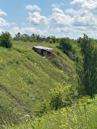 A humble, weathered wooden structure, possibly a shed or small shelter, is situated on a grassy, sloped hillside. The structure has a slightly slanted, corrugated roof and open sides revealing its interior. The surrounding environment is lush with green vegetation, including tall grasses, bushes, and trees. The sky above is blue with scattered white, fluffy clouds. The scene appears to be located in a rural or semi-rural area, consistent with the landscape that could be found in or around the village of Kalinino, Russia. There are no people or animals visible in the image.